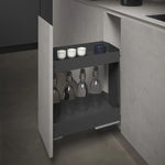 A dark gray under-counter pull-out storage cabinet shown open, holding several glass bottles and small cups, set within a modern kitchen cabinetry interior.